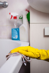 A hand in a yellow glove cleans the bathroom with a multi-colored microfiber cloth against the background of chemicals.