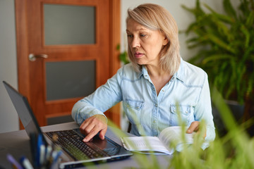 Mature woman looking at laptop at home and reading a book