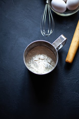 Cooking process. Flour, three eggs in a plate,rolling pin and whisk on a black marble background. Photo for an inscription.