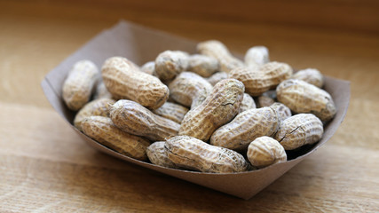 A lot of unpeeled raw peanuts in a brown paper bowl on a wooden table. Delicious and somewhat...