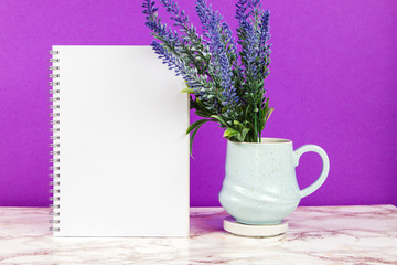 Lavender flowers in a pastel blue mug with blank notebook on a purple and marble background