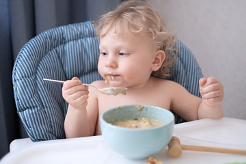 a little boy with blond curly hair sits in a child's chair and eats soup himself with a large spoon from a blue bowl. Eating and educating children on their own