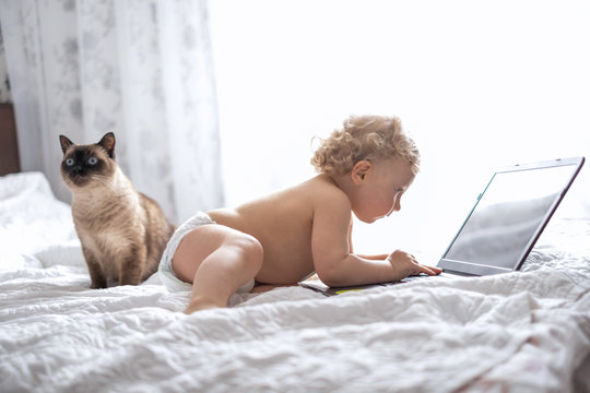 a little boy in a diaper sits on a bed covered with a white blanket and presses buttons on a laptop. Teaching children to work on a computer. Can children sit in front of a computer for a long time?