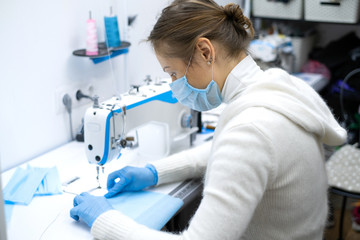 Fototapeta na wymiar a girl in a protective mask sits at the workplace and sew sterile blue masks to protect against the virus. Sewing sterile masks