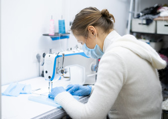 a girl in a protective mask sits at the workplace and sew sterile blue masks to protect against the virus. Sewing sterile masks