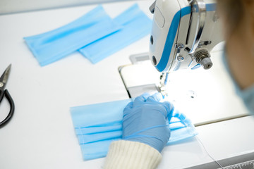 a girl in a protective mask sits at the workplace and sew sterile blue masks to protect against the virus. Sewing sterile masks