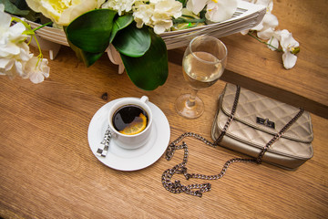 A сup of coffee with lemon and sugar next to a glass of white wine, bouquet of artificial flowers and a woman's bag