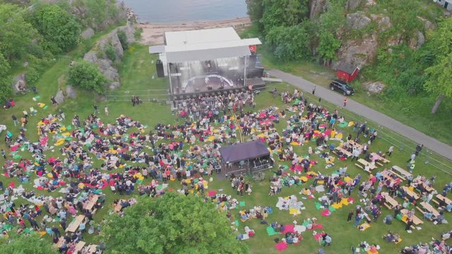 Aerial drone reveal of an outside concert with a substantial amount of people sitting on the grass in a warm summer day.