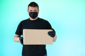 a man with dark hair in a black mask and black sterile gloves holds a cardboard box in his hands....