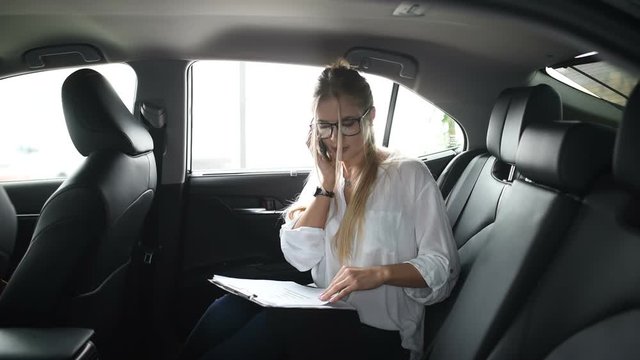 Beautiful business woman is talking on the mobile phone and using a tablet with papers while sitting on back seat in the car.