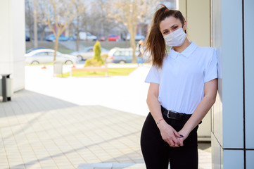 Portrait of a caucasian young brunette girl in a white t-shirt outdoors on a sunny day in the city against the background of a building wall. Model in a medical protective mask.
