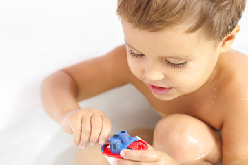 Obraz na płótnie Canvas A happy little boy is sitting in the bathtub and playing with a colorful toy boat in the bathroom. Smiling child in the bathroom on a white background. Children's washing and bathing. Hygiene and