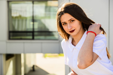 Portrait of caucasian brunette girl in white t-shirt outdoors on a sunny day in the city against the backdrop of the building.