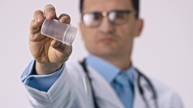 Handsome middle aged male doctor in medical coat, glasses and stethoscope spills down green pills from transparent jar. Close up focused on hand. Impotence of drugs, medicines poisoning, addiction