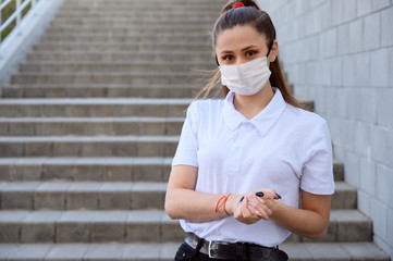 Portrait of a Caucasian young brunette girl in a mask from a virus in a white T-shirt outdoors in the afternoon in the city against the background of a building staircase.