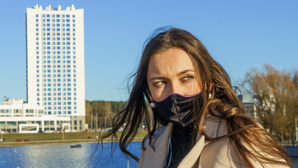 Beautiful young woman in a protective medical mask on the background of the water channel and classic blue sky. Reflection of the sun in water. Covid-19 concept.