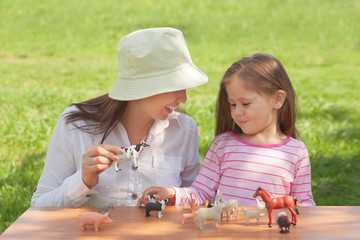 Cute toddler girl and mom play with farm animal figures outdoors. Summer vacation childhood in the countryside. The child learns farm animals. Early education and development