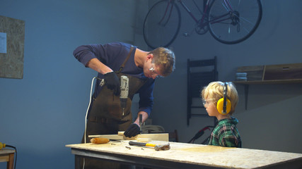 Boy watches his dad drill a board. Medium long shot