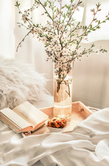 The atmosphere of a romantic morning, breakfast in bed. Flowering branches in a vase, an open book and a biscuit on a wooden tray on a white bed. Home interior. Lifestyle