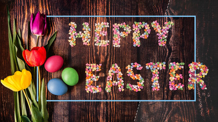 
Easter composition of colorful tulips with colored Easter eggs and the inscription Happy Easter from colored sugar sprinkles on a wooden table.