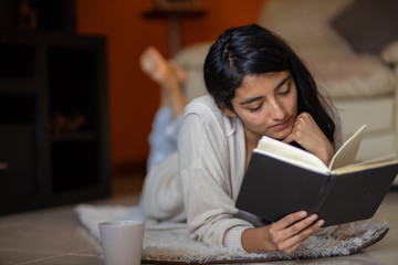 woman latina  lying down looking at the side with book with cup of coffee in pandemic house,...