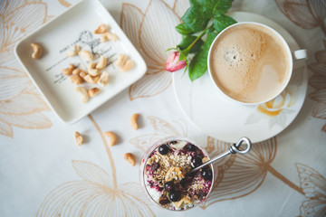 Top view of a tablecloth covered with a bowl of organic cottage cheese yogurt with granola and black currant, a garden rose, a plate with cashew nuts and a cup of delicious morning coffee. Toning