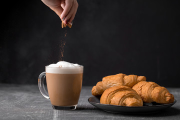 Side view on cappuccino coffee with milk foam sprinkled with croissants in a transparent glass mug with with a hand pouring ground cinnamon on a gray background, horizontal format - Powered by Adobe