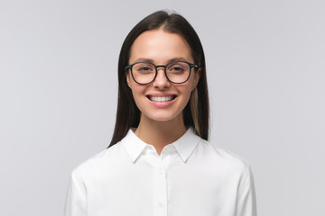 Smiling female in eyeglasses wearing white collar shirt, looking at viewers with happy smile, feeling confident and ready to help, isolated on gray background