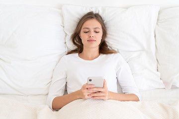 Young woman lying in bed on pillows, wearing white clothes, holding smartphone with both hands and reading posts or messages after awakening in the morning