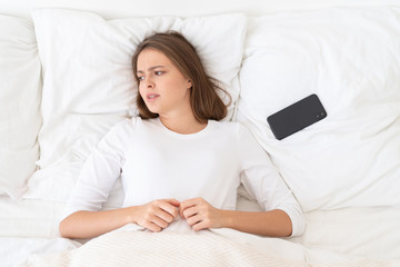 Obraz na płótnie Canvas Young female put away her smartphone after breakup with lover, lying in bed in morning, looking helpless, stressed and lonely