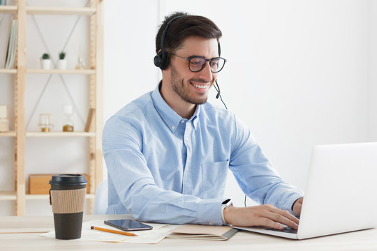 Young Man Working In Hotline Answering Client Inquiries And Helping Solve Problems, Feeling Positive And Confident, Smiling Happily, Typing Data On Laptop In Office