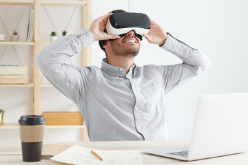 Young IT guy wearing gray shirt and virtual reality headset, sitting in office and experiencing VR opportunities of 3D gaming, smiling happily
