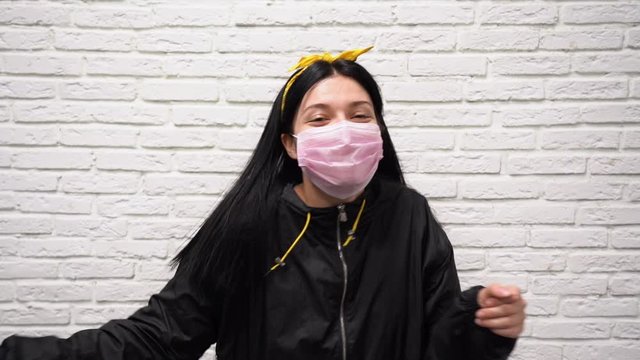 Pretty happy Caucasian woman with long black hair in sport jacket and a yellow headband in pink protective medical mask on her face dance over the background of a white brick wall slow motion close up