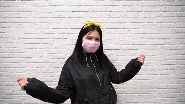 Attractive happy Caucasian girl with long black hair in a sport jacket and yellow headband in pink medical disposable filter mask dance against the background of a white brick wall slow motion.