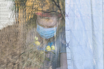 A man in medical mask near the window looking at blooming rose.