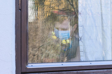A man in medical mask near the window smelling blooming rose.