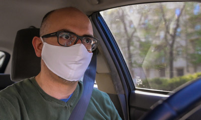 Turin, Piedmont, Italy. April 2020. Coronavirus pandemic: portrait of a Caucasian man driving the car wearing a white mask to avoid contagion. Selective focus on man and blurred background.