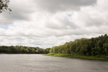 Dark and light parts. River running across with reflections in the water. Grey sky. Green forest on the other side