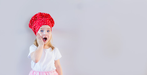 Little blonde smiling girl in red cook's hat staying on neutral grey background and holding her cheek, surpised, shocked. Space for text