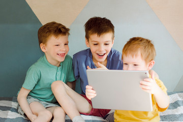 three children are sitting on the bed in the children's room, looking at the tablet screen and laughing