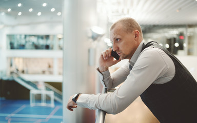 A serious robust hispanic man entrepreneur is having a phone conversation while leaning against chrome railing of a balcony in a contemporary office interior, with a copy space place on the left