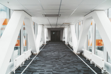 Bright long modern passageway between two towers of an office or a hospital skyscraper with  zig-zag lines of the load-bearing beams, suspended ceiling, heating batteries, and carpeting on the floor - Powered by Adobe