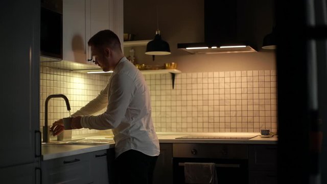 Handsome bearded man filling glass with tap water in modern dark kitchen and drinks it. Thirst guy drinking water. Shotting in slow motion.