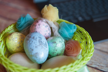 Colorful Easter eggs, decorative decoration flowers lie in a bright green wicker basket on a wooden table. Laptop, work from home. Working on holidays