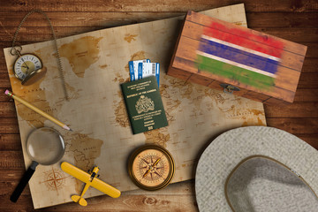 Top view of traveling gadgets, vintage map, magnify glass, hat and airplane model on the wood table background. On center, official passport of Gambia and your flag.