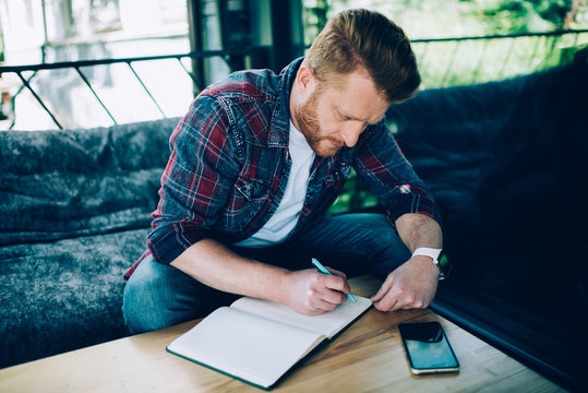 Concentrated guy sitting at table and writing in notebook