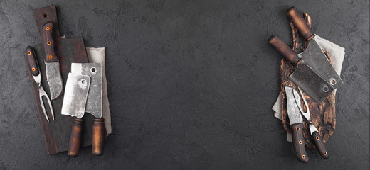 Vintage chopping board with meat hatchets and fork with knife on black background with salt and pepper.