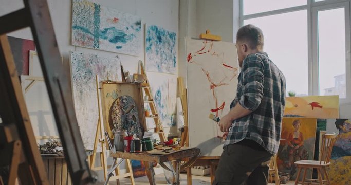 Rear of talanted young Caucasian male artist painting on canvas with red paint and brush in workshop. Back view on man with beard working in artistic studio at easel in daytime. Fine art concept.