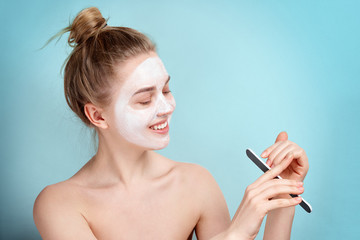 Obraz premium Young girl in a cosmetic mask of white smiling face. Skincare. Blonde woman smiling on blue background. moisturizer mask.