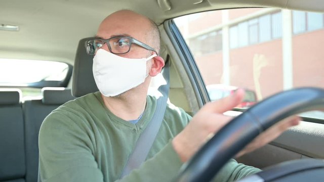 Turin, Piedmont, Italy. April 2020. Coronavirus pandemic: portrait of a Caucasian man driving the car wearing a white mask to avoid contagion. Selective focus on man and blurred background.